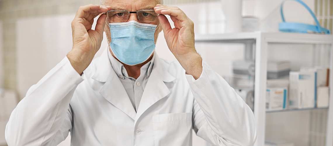 Man in lab coat wearing a surgical mask adjusts glasses in a lab.