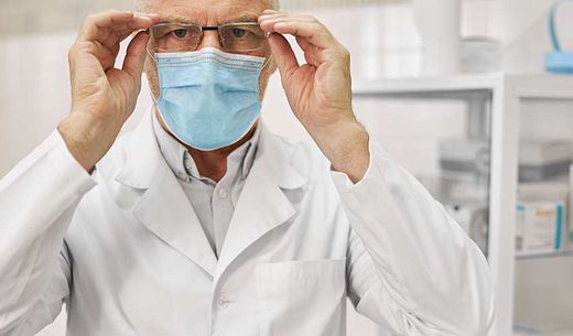 Man in lab coat wearing a surgical mask adjusts glasses in a lab.
