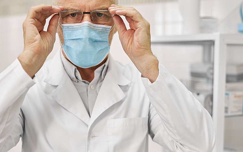 Man in lab coat wearing a surgical mask adjusts glasses in a lab.