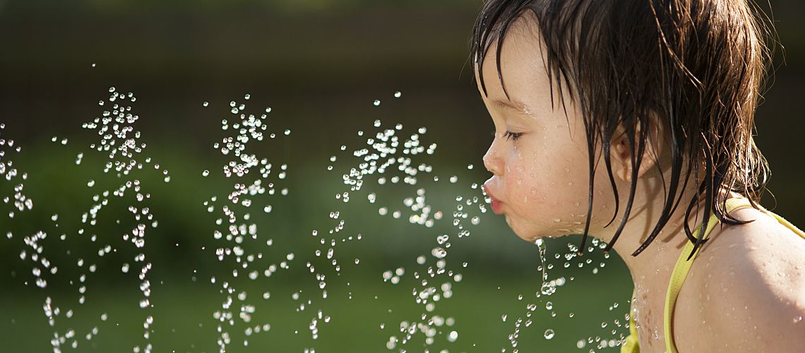 Young toddler drinking from a water sprinkler in her backyard.