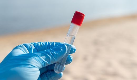 A gloved hand holds a water sample on a river beach.