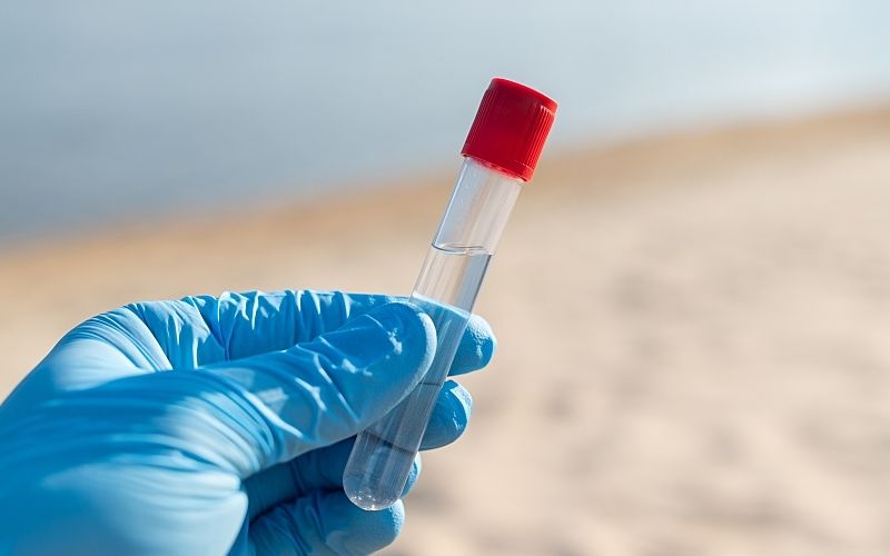 A gloved hand holds a water sample on a river beach.