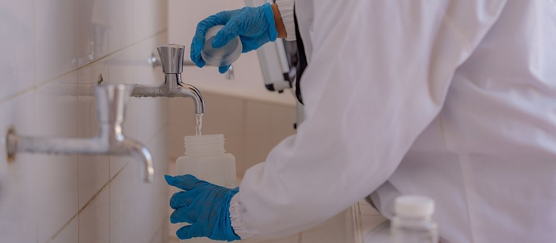 A gloved lab worker takes a drinking water sample from a tiled sink.
