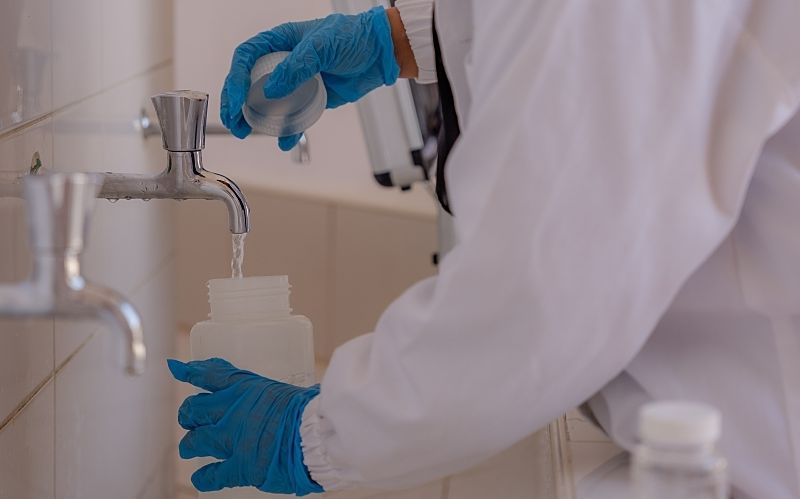 A gloved lab worker takes a drinking water sample from a tiled sink.