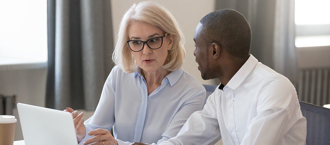 A manager sits with a younger employee at a laptop.