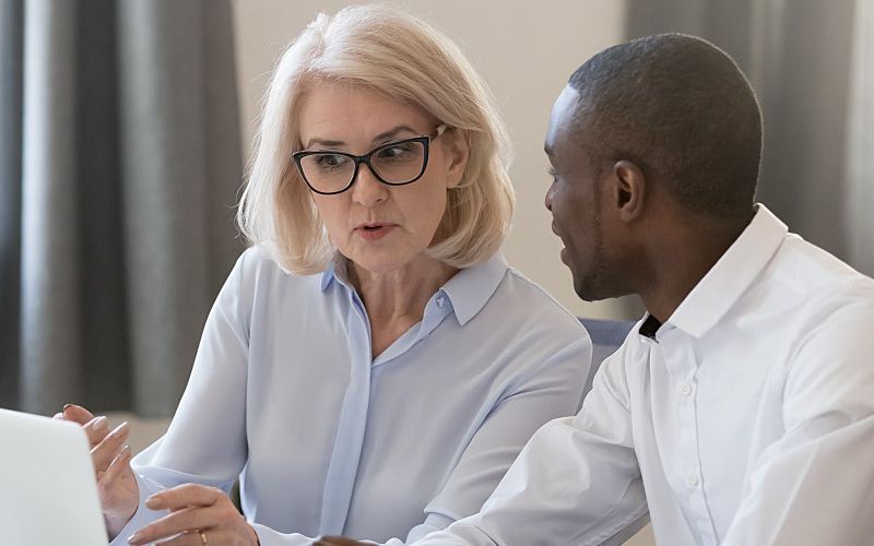 A manager sits with a younger employee at a laptop.