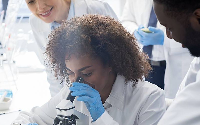 Three scientists observe a lab sample through a microscope.