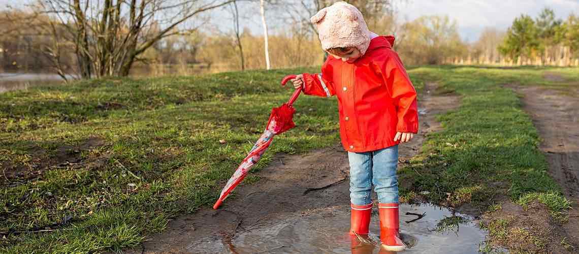 A child in a red rain slicker stands in a puddle at the park.