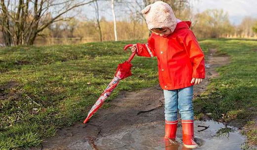A child in a red rain slicker stands in a puddle at the park.