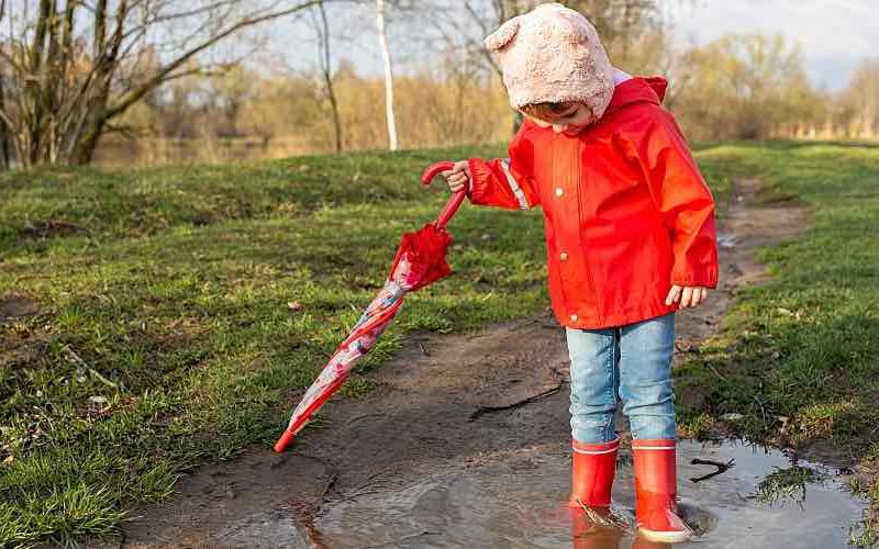 A child in a red rain slicker stands in a puddle at the park.