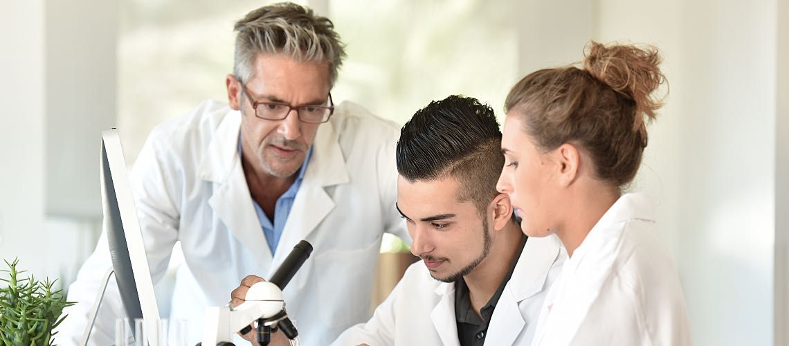 A lab manager instructs two team members sitting at a microscope.