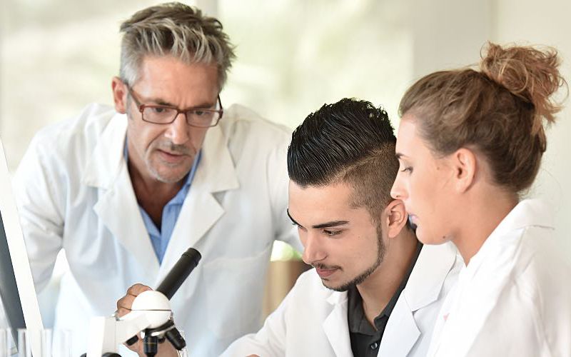 A lab manager instructs two team members sitting at a microscope.