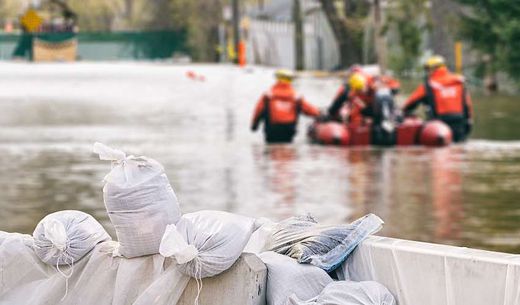 Rescuers wade through water on a flooded street with sandbags in the foreground.