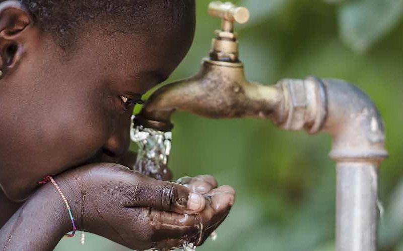 A child drinking water from a tap in a leafy environment.