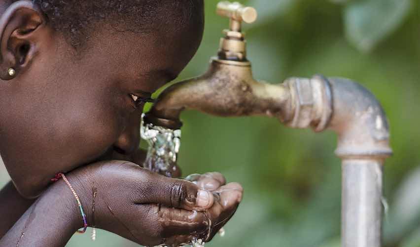 A child drinking water from a tap in a leafy environment.